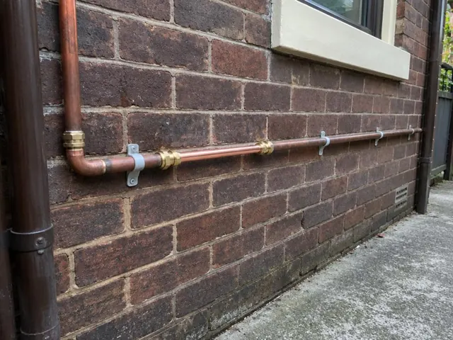 Copper water pipe run on a brick exterior wall beside a window in Ashfield